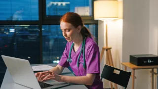 A women in scrubs sits at her desk while typing on a her laptop