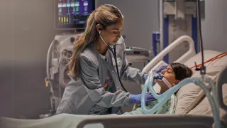 A girl lies in a hospital bed while a healthcare professional attends to her. There is a PrisMax device in the background.
