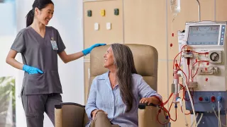 A woman in a chair is connected to the AK 98 Dialysis Machine, and conversing with a nurse standing beside her