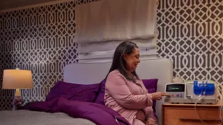A woman is sitting on her bed in her bedroom, interacting with her Homechoice Claria device