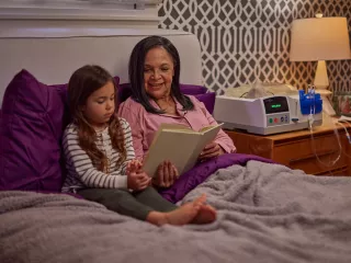 A grandmother and her granddaughter are reading together in bed. A Homechoice Claria device sits on the nightstand 