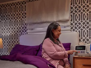 A woman is sitting on her bed in her bedroom, interacting with her Homechoice Claria device